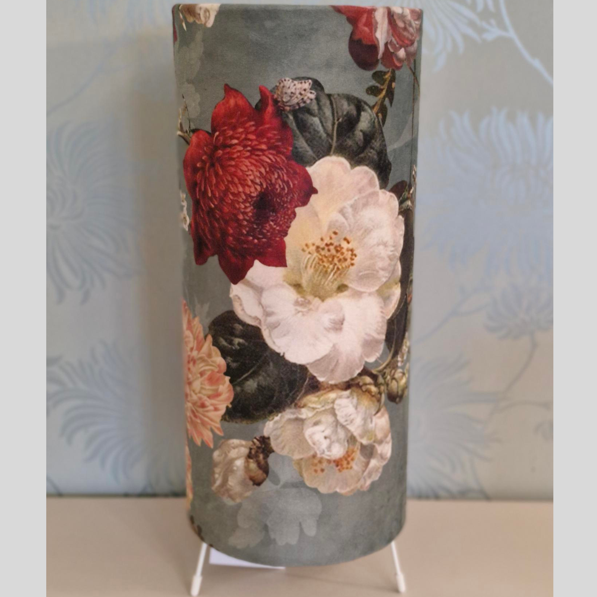 A cylindrical lampshade featuring a vintage floral print with large red, white, and peach flowers on a muted gray-blue background, displayed on a small white stand.