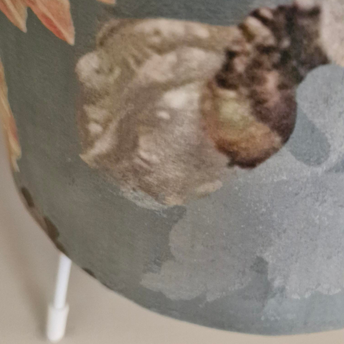 Close-up of a lampshade featuring a textured green-gray base with a printed botanical pattern in muted green, brown, and peach tones, with a small white support leg visible at the bottom.